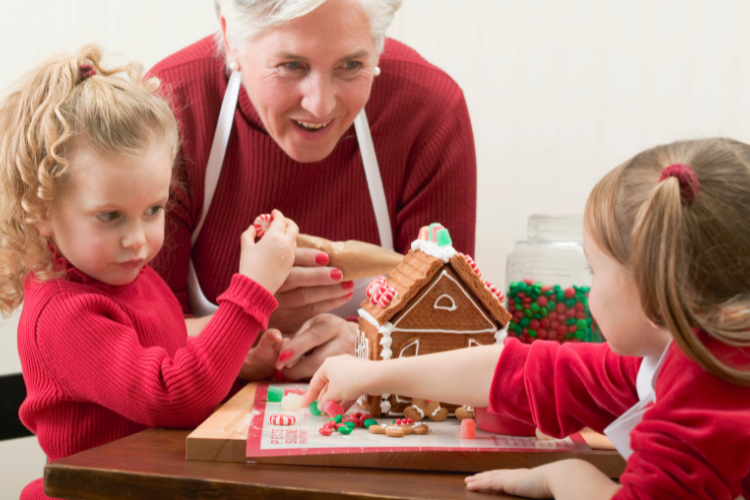 Two children decorating a gingerbread house with their grandmother