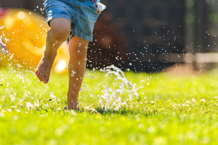Close up of child's legs running through sprinkler on green grass