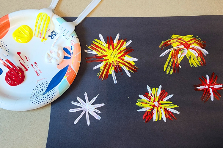 A plate of red, yellow, and white paint next to a black piece of paper with painted fireworks.