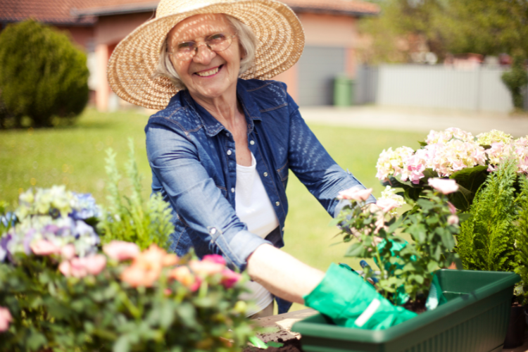 A woman in a wide brim hat and gardening gloves smiles as she plants flowers.