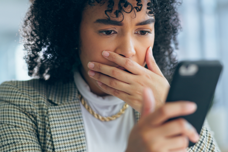 A horrified woman looking at her cell phone while covering her mouth with her hand.